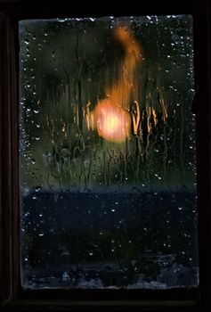 A window covered in raindrops with a blurry, illuminated background creating a moody atmosphere.
