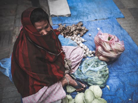 A woman selling vegetables at a street market, wrapped in a red scarf, sitting on a blue mat.