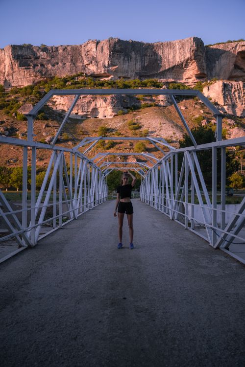 Free A woman poses confidently on a metal footbridge against a rocky landscape backdrop during sunset. Stock Photo