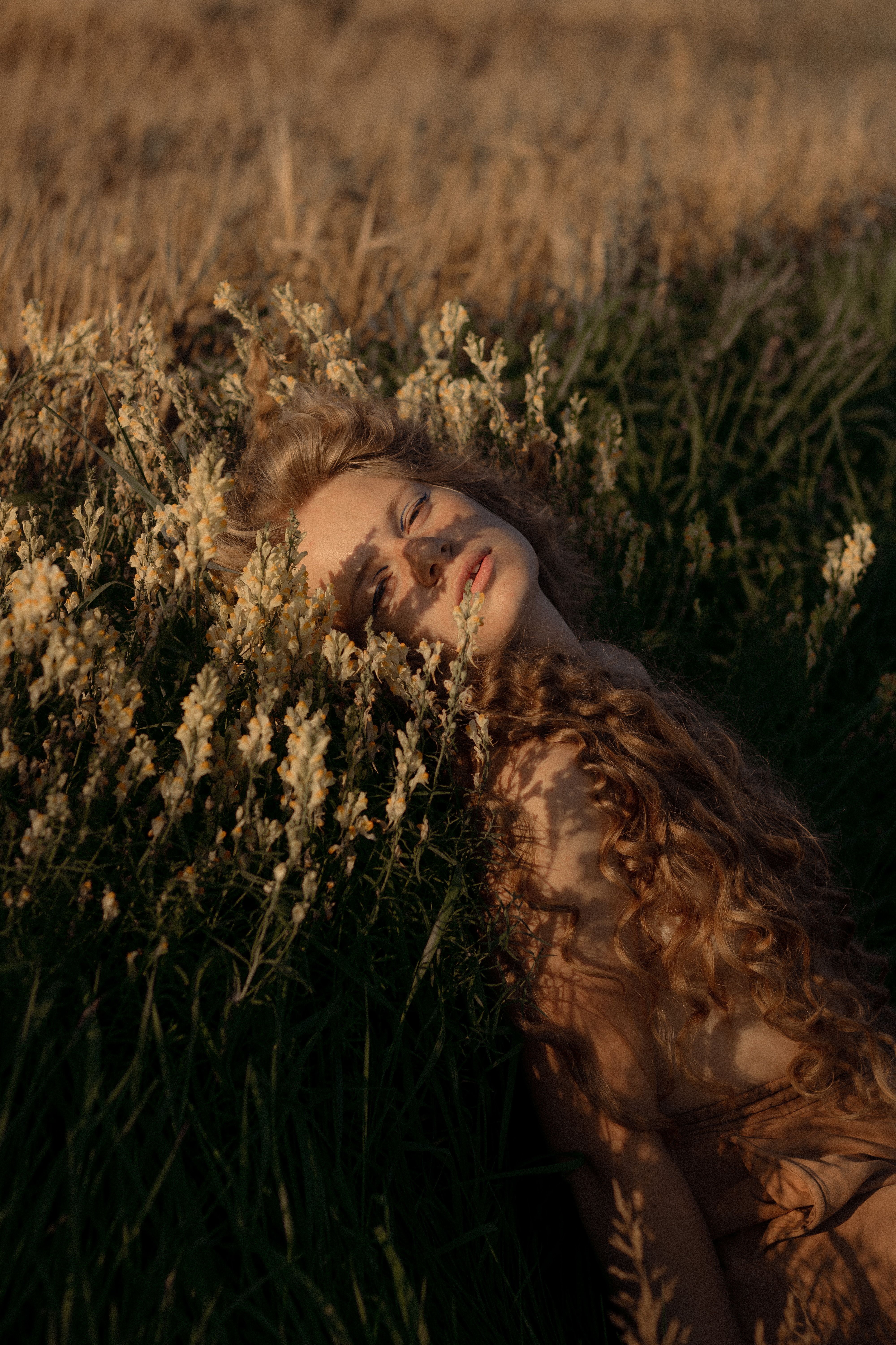 A woman with curly hair lying in a sunlit field, surrounded by tall grass and flowers.