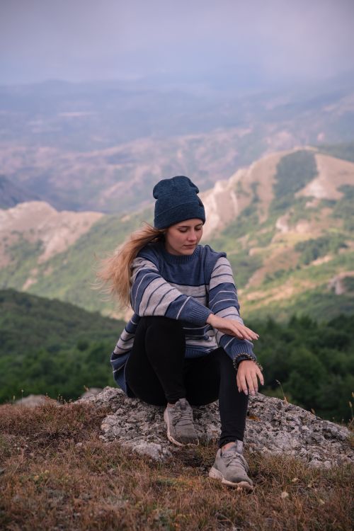 Free Woman wearing a beanie and sweater sits on a mountain, enjoying the scenic view. Stock Photo