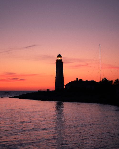 Free Lighthouse silhouetted by a vivid sunset over a calm waterfront, evoking serenity and navigation. Stock Photo
