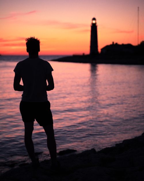 Free Silhouette of a person by a lighthouse with vibrant sunset along the seashore. Stock Photo