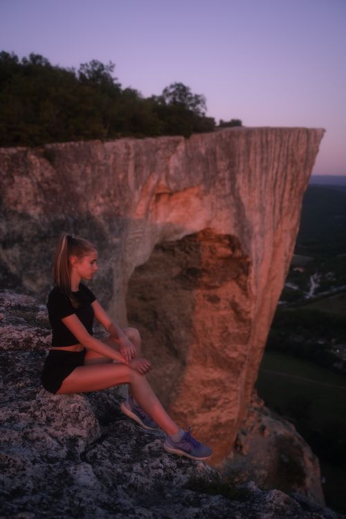 Free Young woman sitting on a cliff edge, enjoying a scenic sunset view with rocky formations. Stock Photo