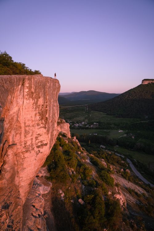 Free Silhouette of a person standing on a steep cliff overlooking vast landscape during a stunning sunset. Stock Photo