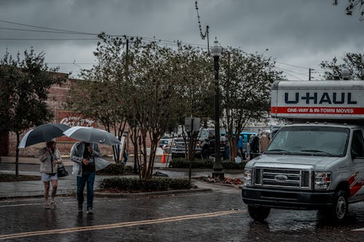 People with umbrellas walking by U-Haul truck on a rainy city street.