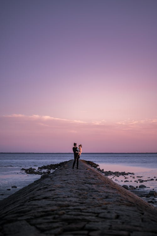 Woman Holding Her Daughter and Standing on the Seashore at Sunset 