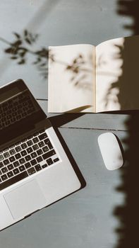 A sunlit workspace featuring a laptop, open notebook, and wireless mouse casting artistic shadows.