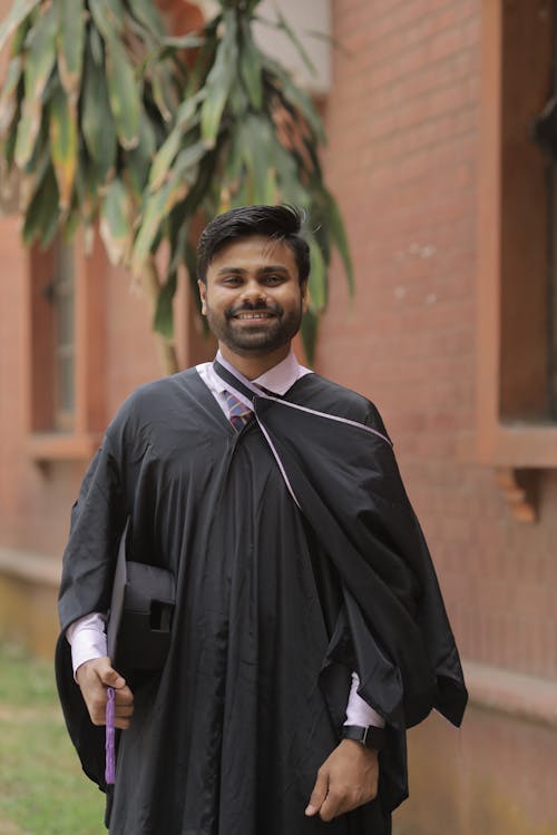 A Man in Black Academic Dress Standing Near Brick Building while Smiling at the Camera