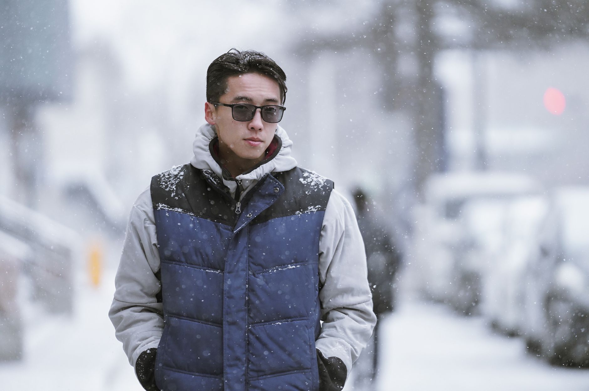 Asian man in sunglasses walking through a snowy urban street, displaying winter fashion.