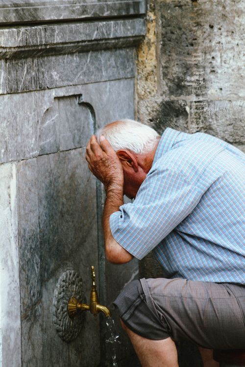 Free Senior man in casual attire washes face at an outdoor tap, emphasizing refreshment and daily life. Stock Photo