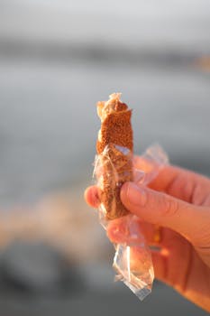 Close-up of a hand holding a wrapped brown sugar candy with a blurred oceanic background, capturing a sweet moment.