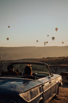 A vintage convertible overlooks a breathtaking landscape with hot air balloons in Cappadocia, Türkiye.
