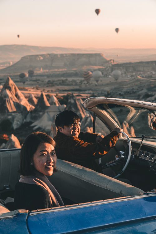 Free A couple in a classic car enjoying the Cappadocia landscape with hot air balloons at sunrise in Türkiye. Stock Photo