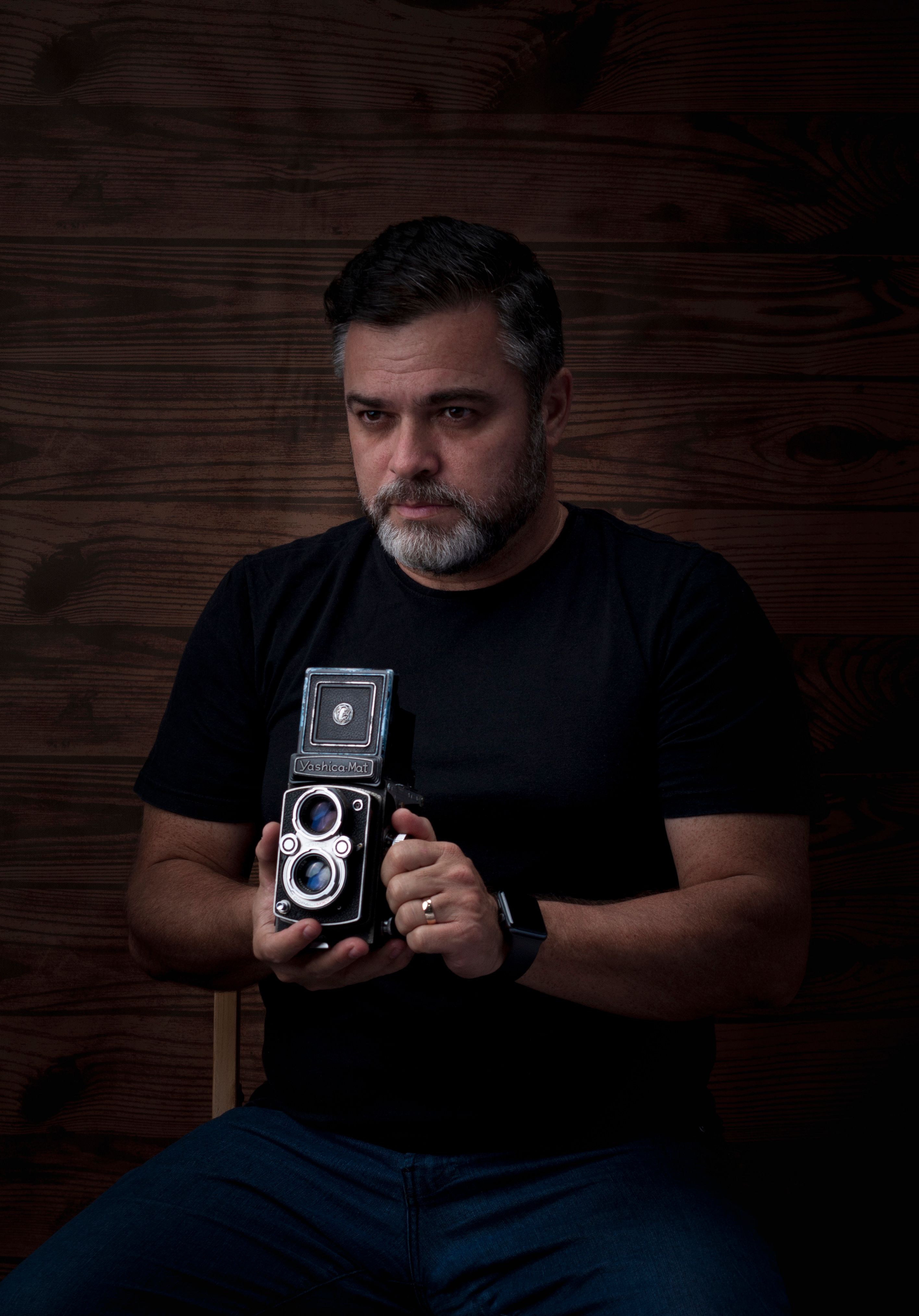 Stoic man with beard holding a vintage twin lens camera against wooden background.