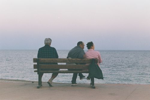 Free Three people sit together on a bench by the tranquil seaside during sunset, enjoying a serene moment. Stock Photo