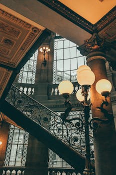 A classic vintage interior featuring ornate stairs, windows, and decorative lamps.
