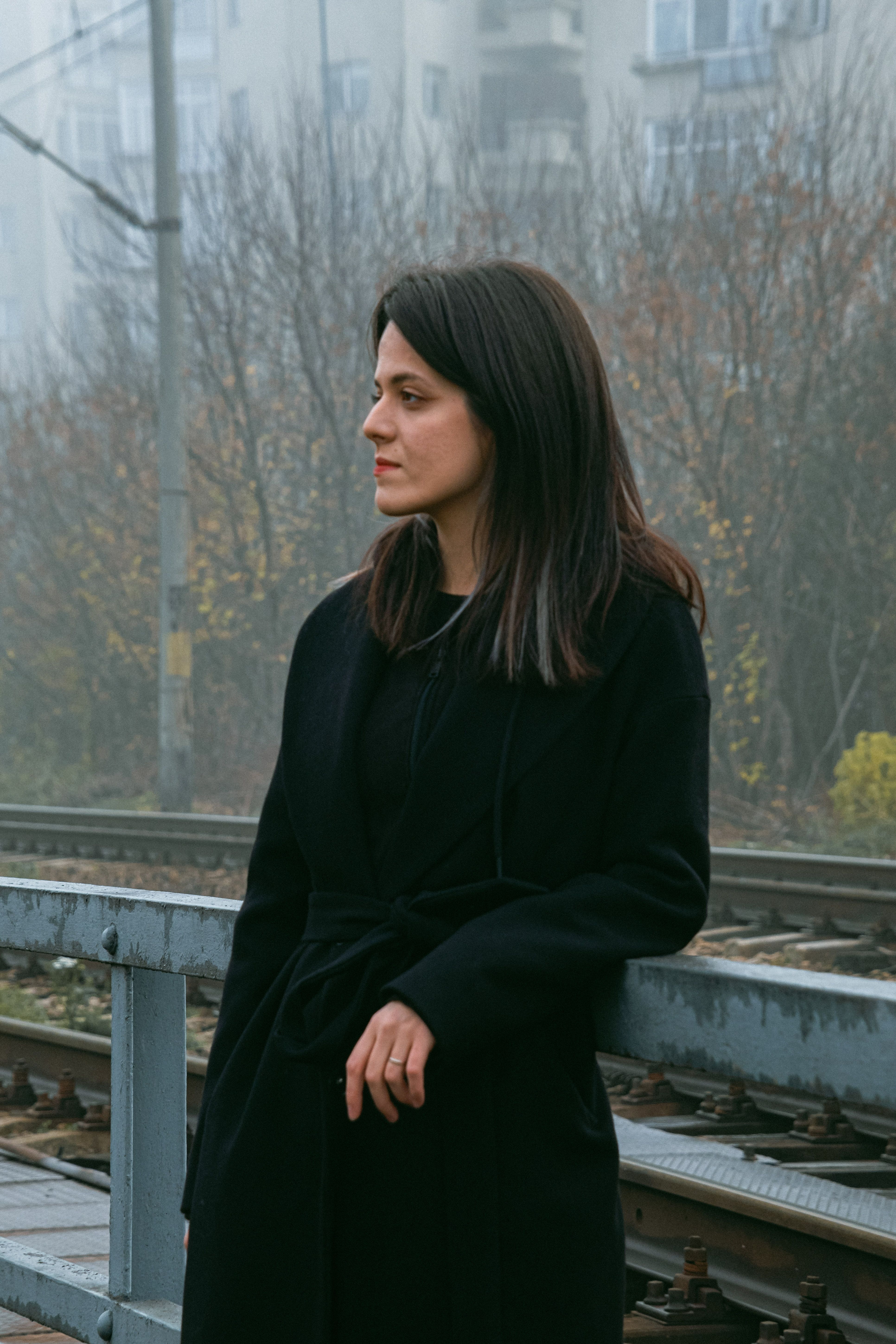 A woman in a black coat standing thoughtfully by railway tracks on a foggy day.