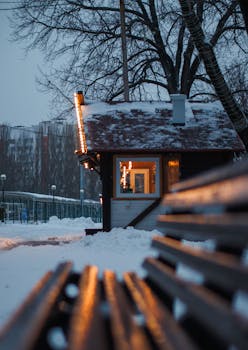 A cozy cabin surrounded by snow in a winter urban setting with a wooden bench in the foreground.