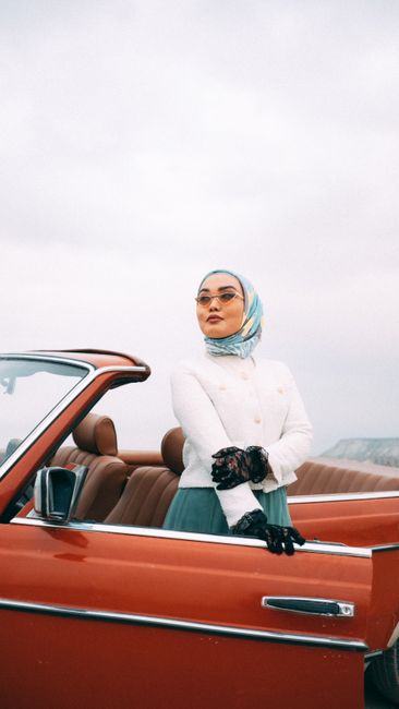 Confident woman in hijab poses next to a red convertible under overcast skies.