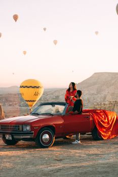 Couple enjoys sunrise in Cappadocia with hot air balloons and red car.