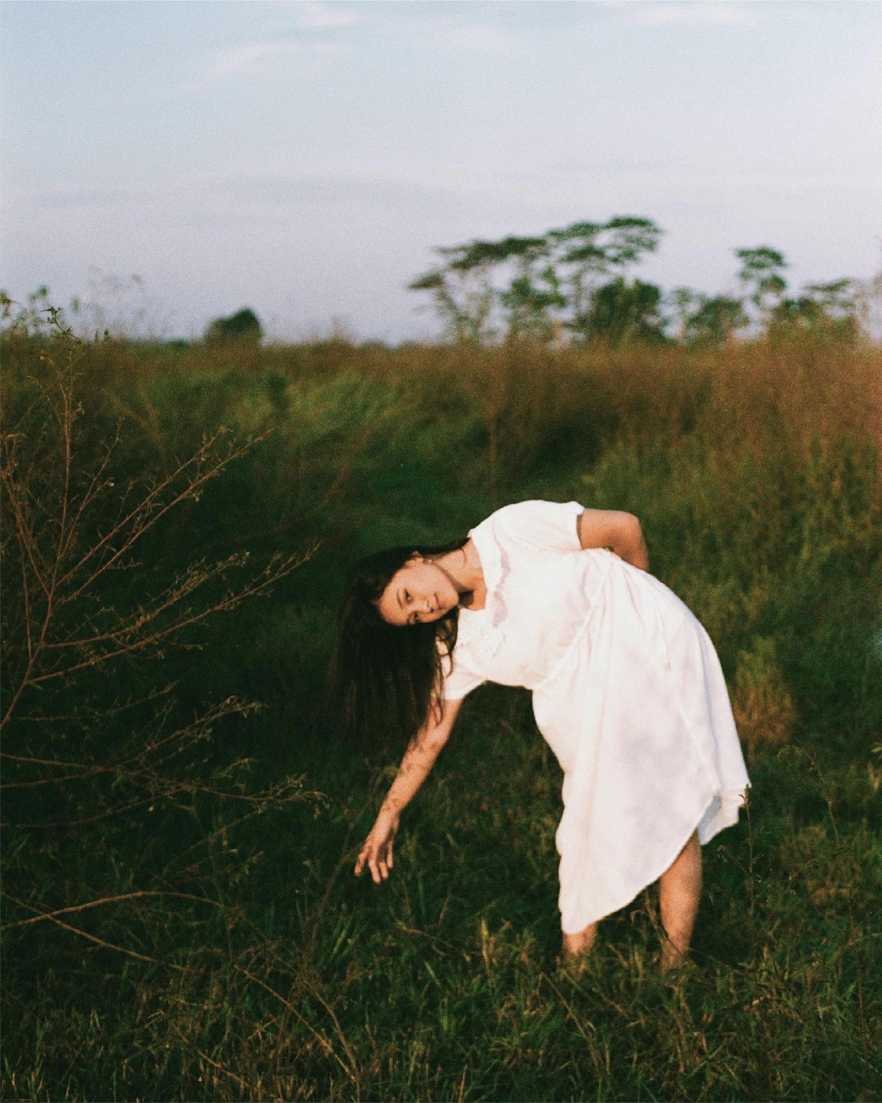 A woman in a white dress poses gracefully in a lush grass field during daytime.