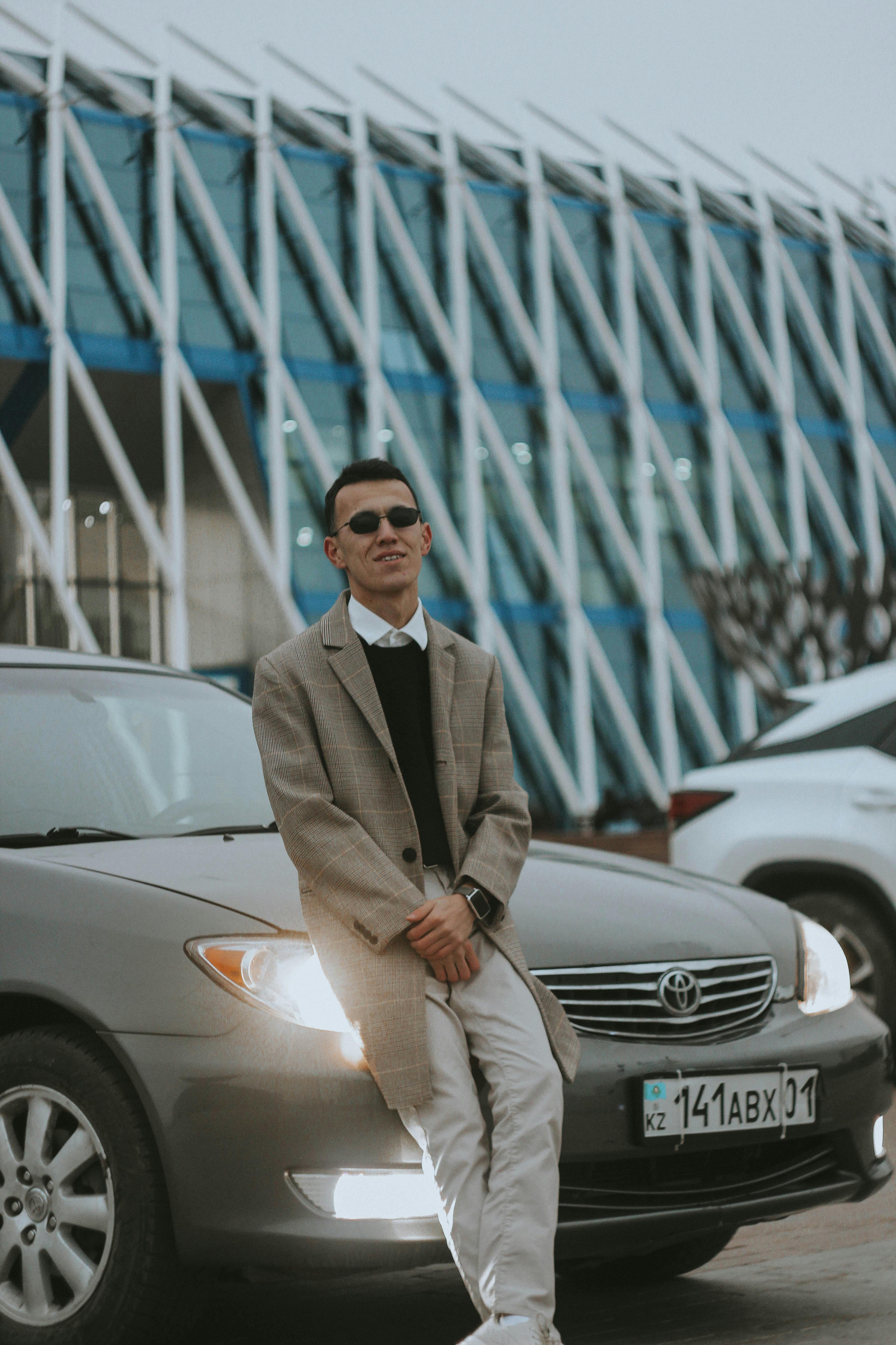 A fashionably dressed young man with sunglasses leans on a car in a city environment.