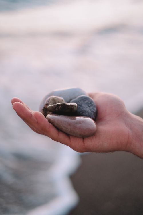 Free A hand gently cradling pebbles against a blurred ocean background. Stock Photo