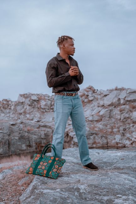 Man in stylish outfit posing on rocky landscape with bag, showcasing fashion outdoors.
