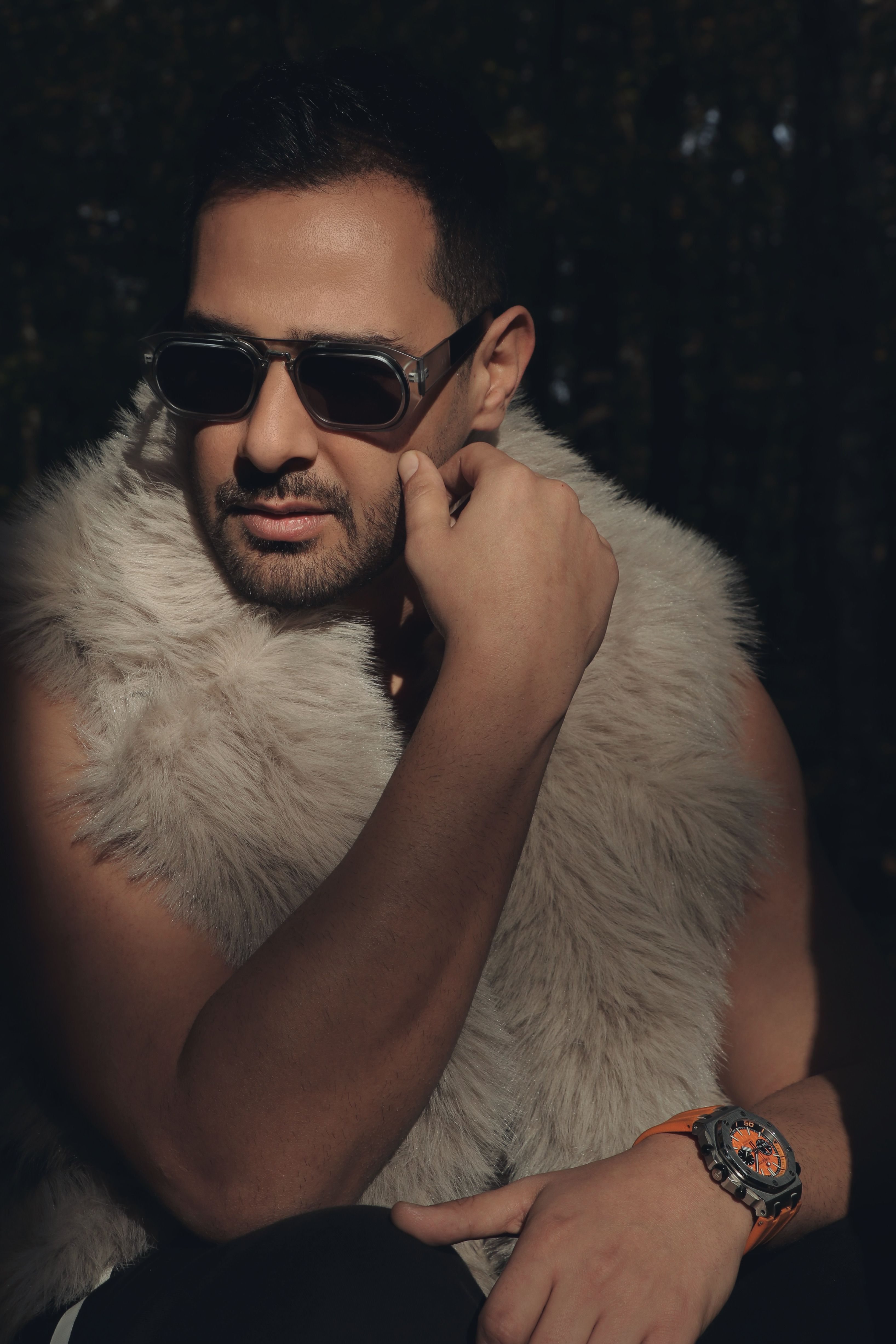 A fashionable man wearing sunglasses and a fur vest posing confidently outdoors.