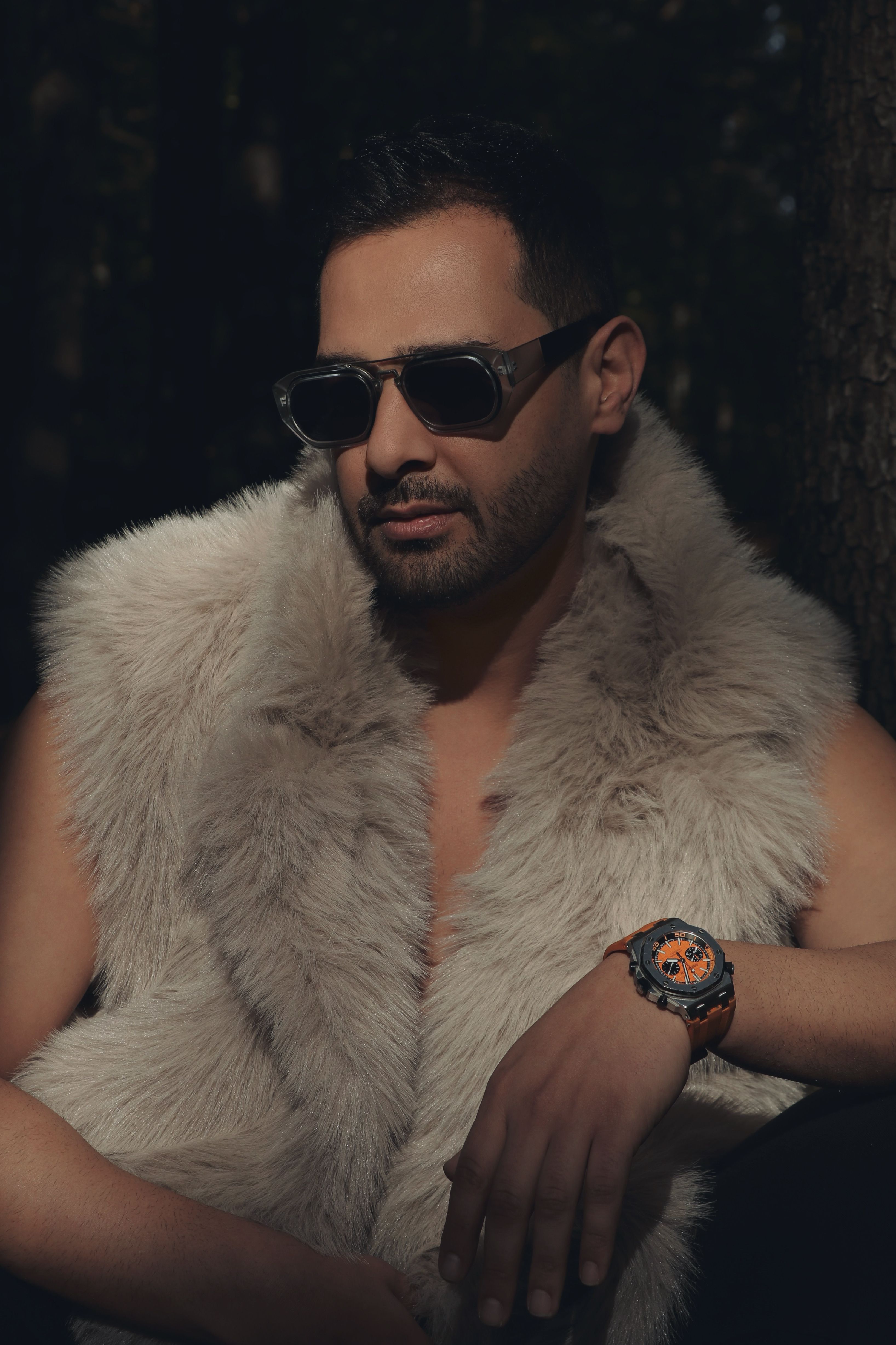 Confident man in a fur vest and sunglasses poses stylishly outdoors.