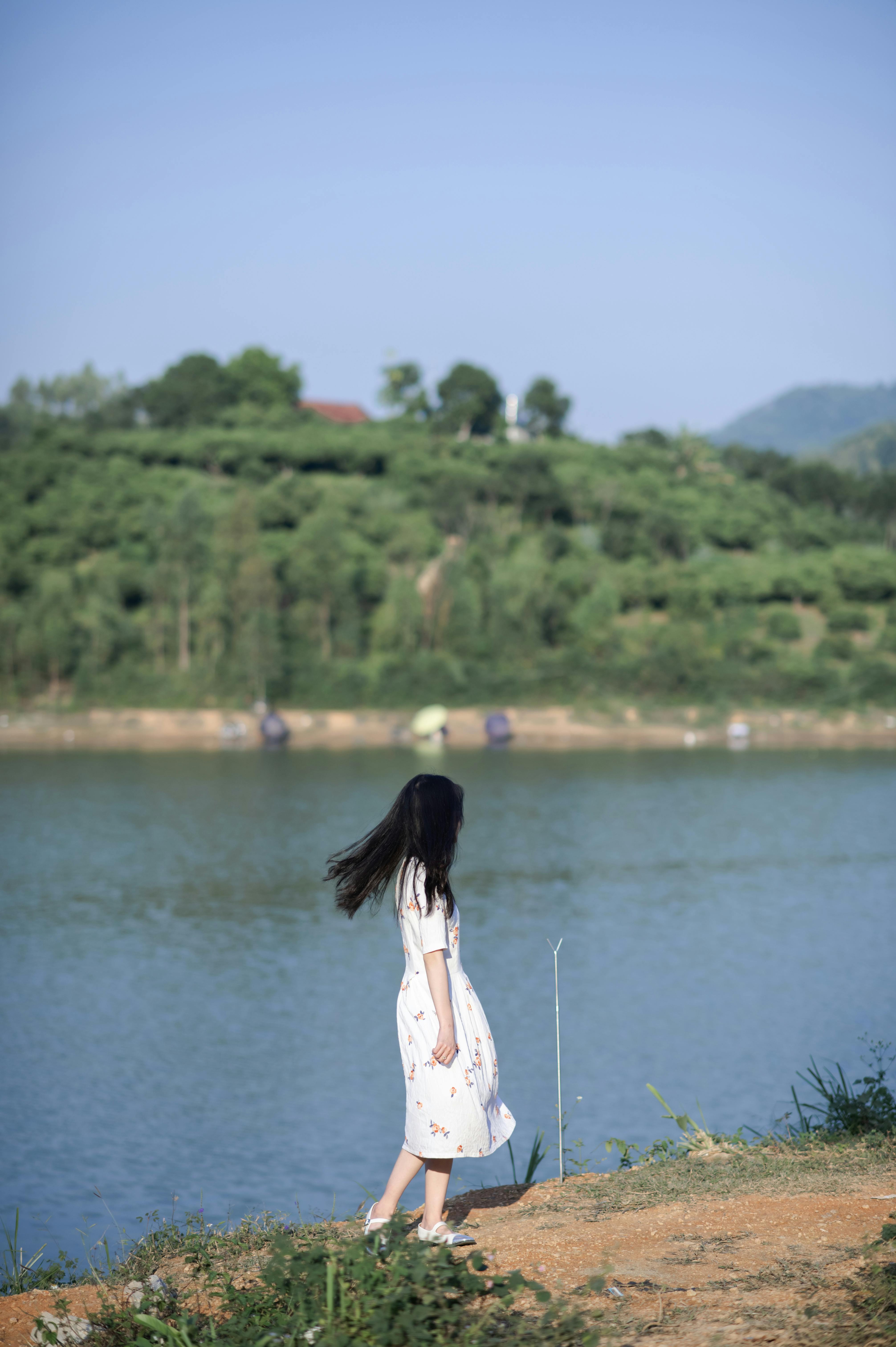 A serene scene of a woman in a white dress walking by a lake surrounded by lush greenery.