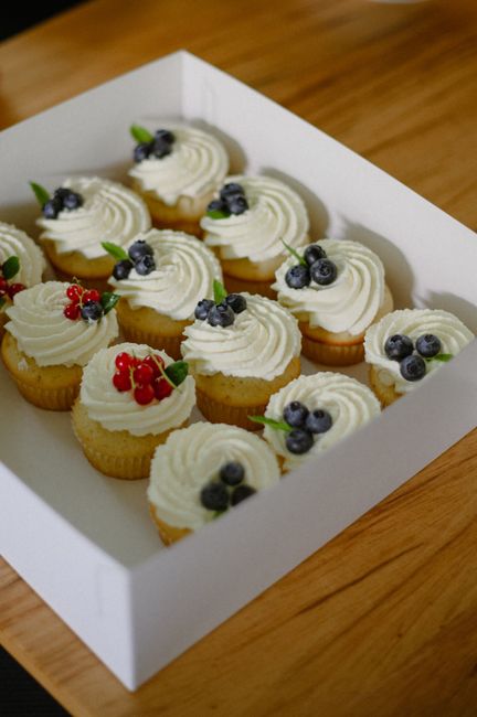 Twelve vanilla cupcakes topped with cream and fresh berries in a white box on a wooden table.