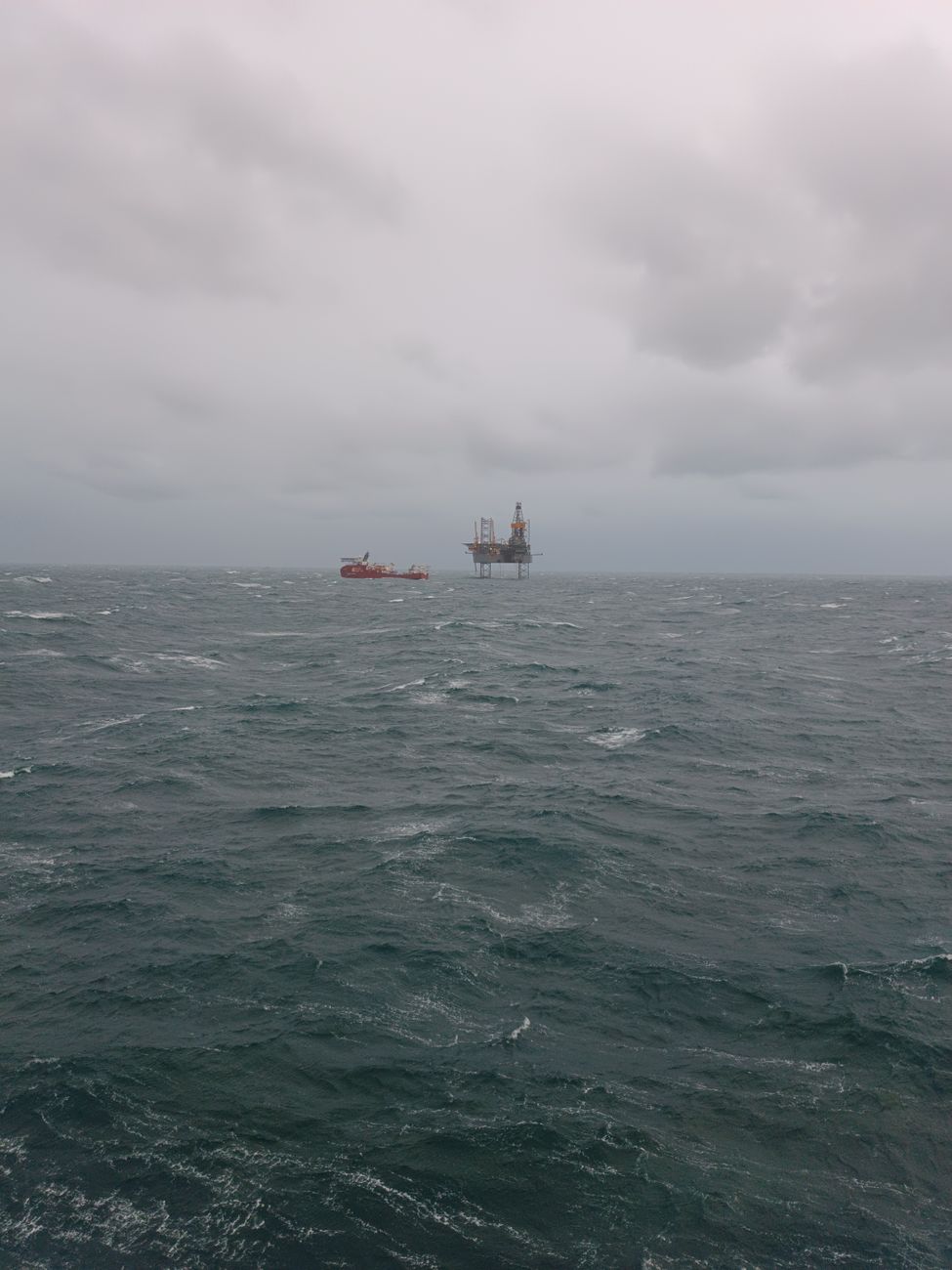 Een verre blik op een offshore olieboorplatform en bevoorradingsschip te midden van stormachtige zeeën onder bewolkte luchten.