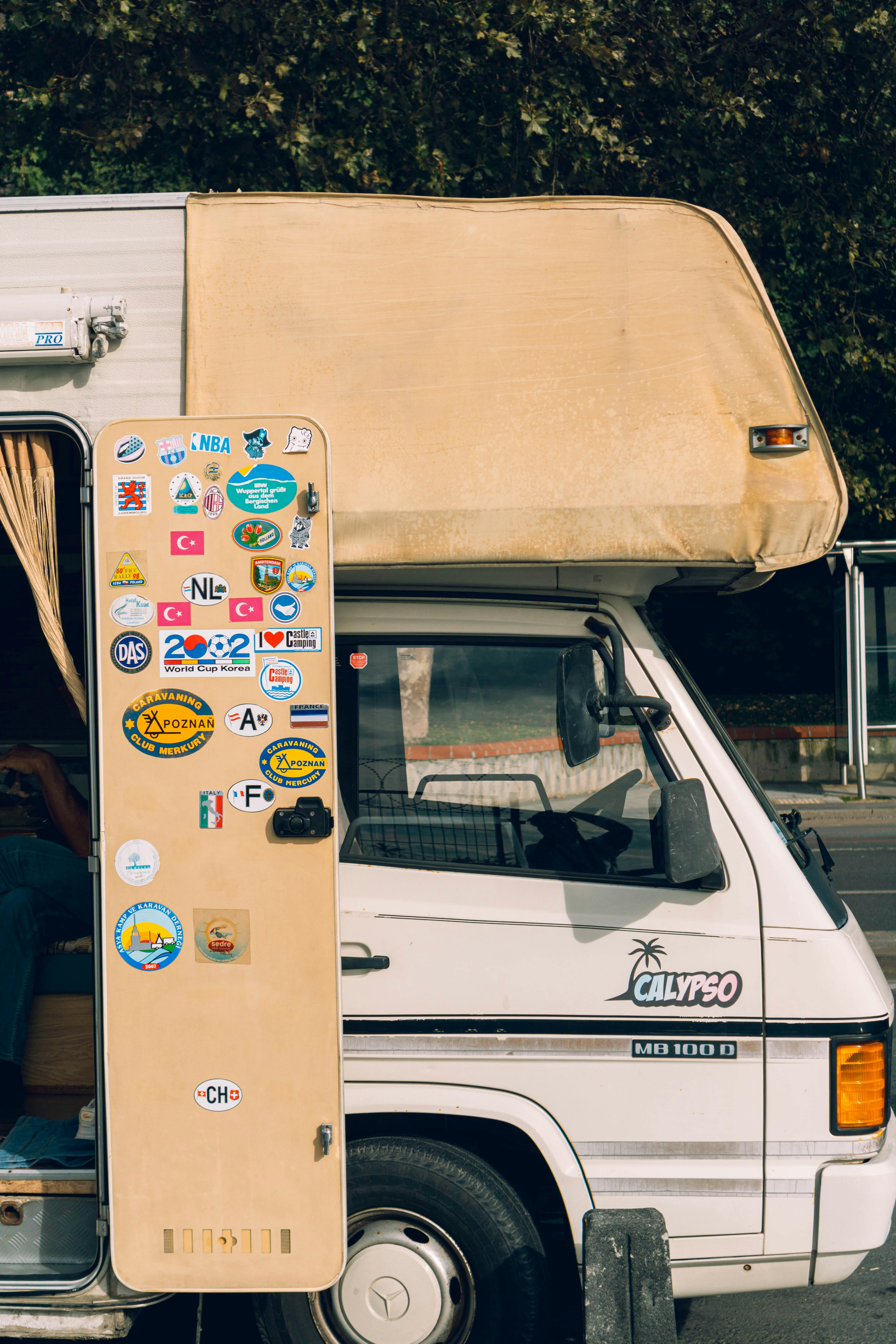 A classic campervan adorned with travel stickers parked outdoors under leafy trees.