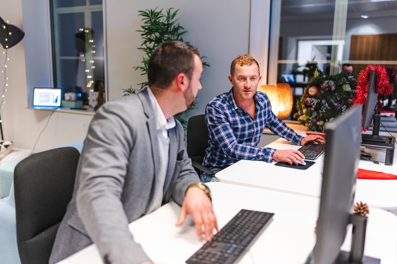 Two men collaborate in a modern office with computers and holiday decor.