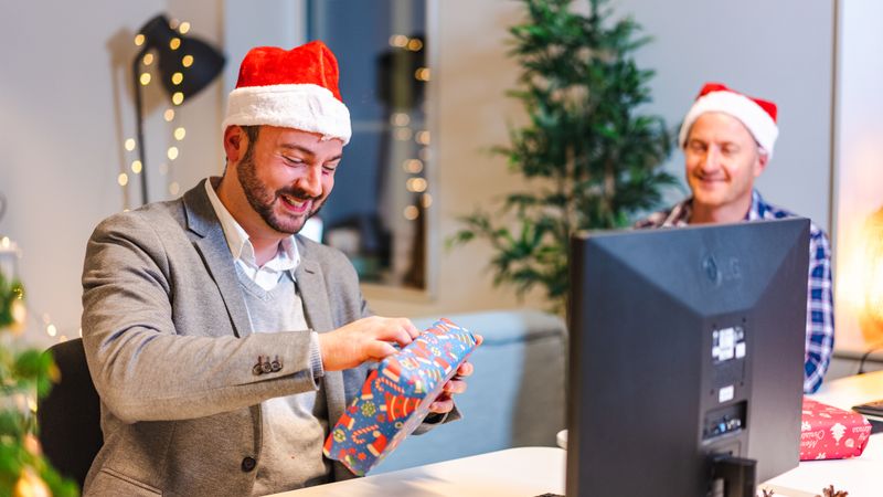 Zwei Kollegen mit roten Weihnachtsmützen im Büro, einer packt am Schreibtisch lachend ein bunt verpacktes Geschenk aus