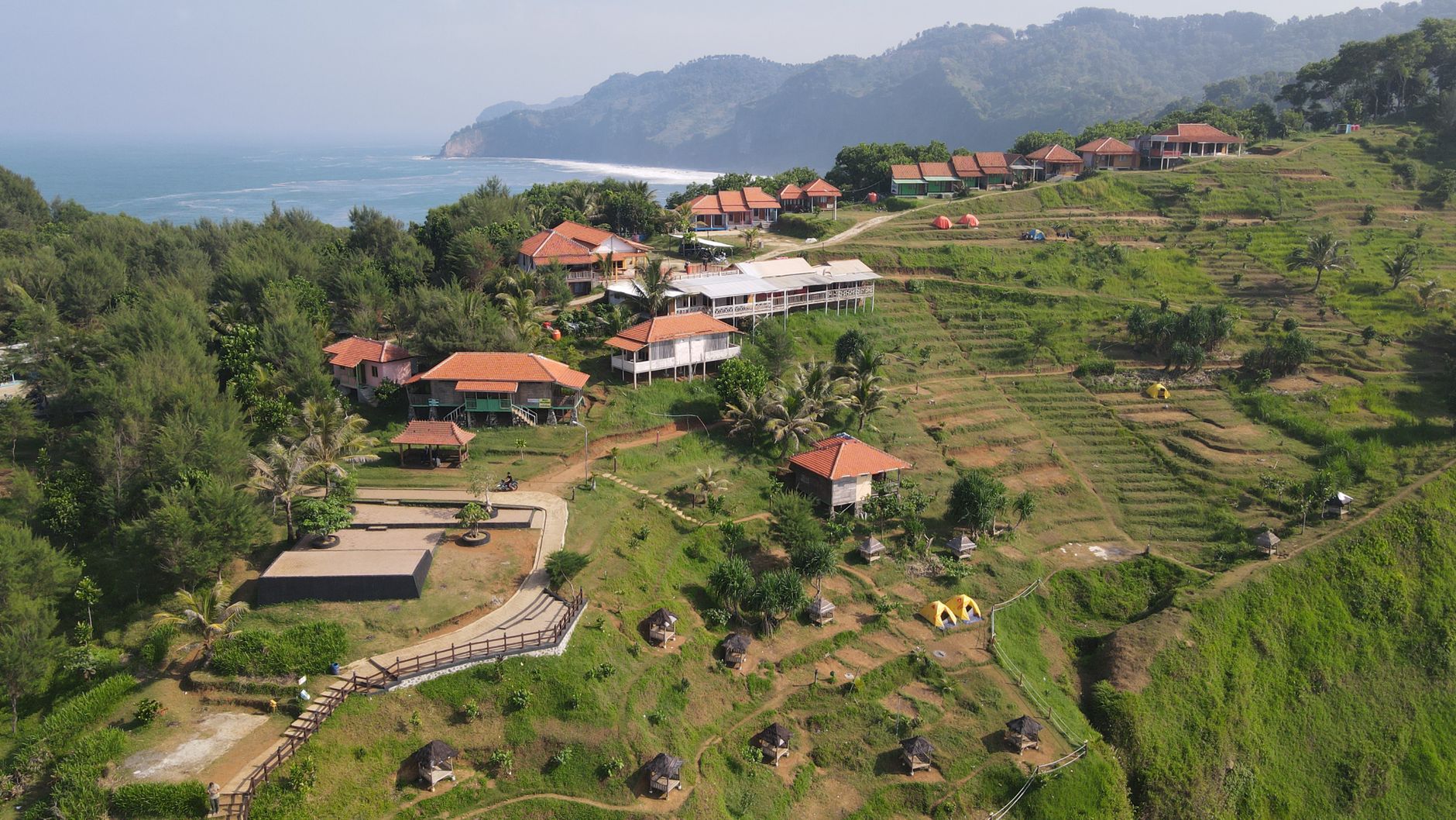 Breathtaking aerial shot of a coastal village in Kebumen, Indonesia, featuring lush green hills and red-roofed houses by the sea.
