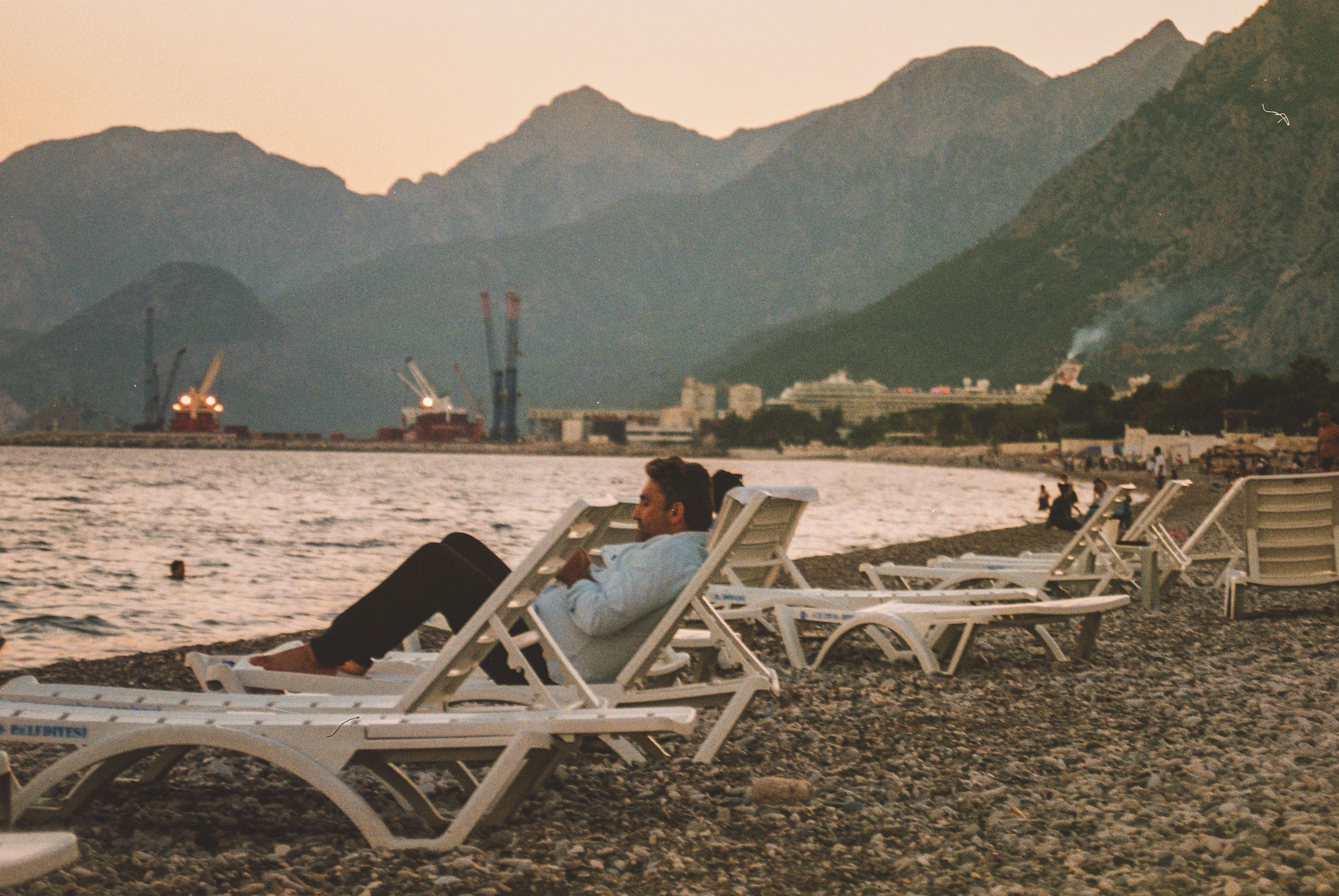 A man relaxes on a beach chair at sunset, with mountains in the background.