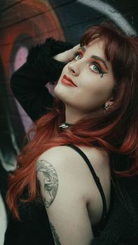 Redheaded woman with striking eye makeup posing provocatively against a graffiti wall.