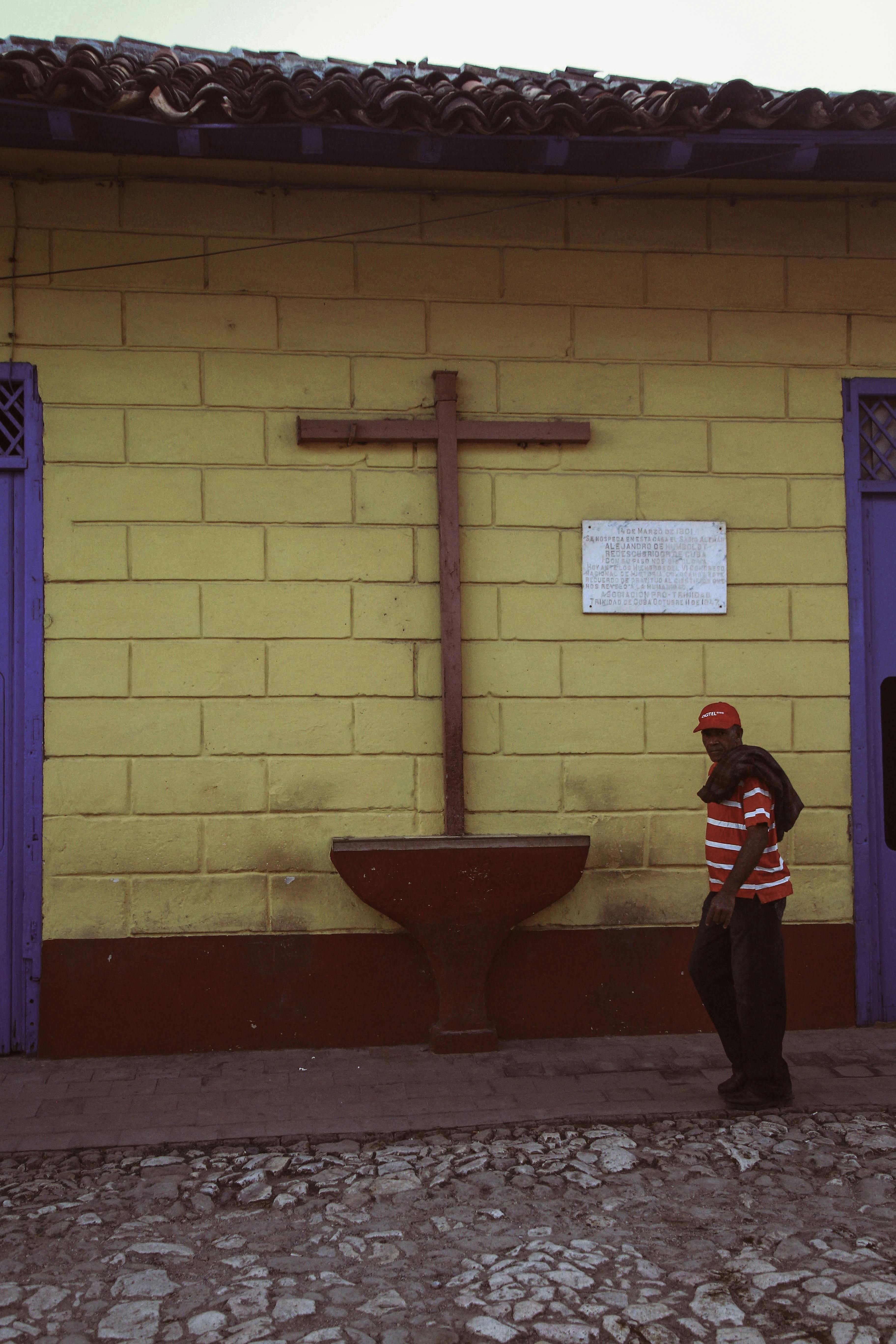 A man in a striped shirt walks past a rustic wall featuring a wooden cross and plaque.
