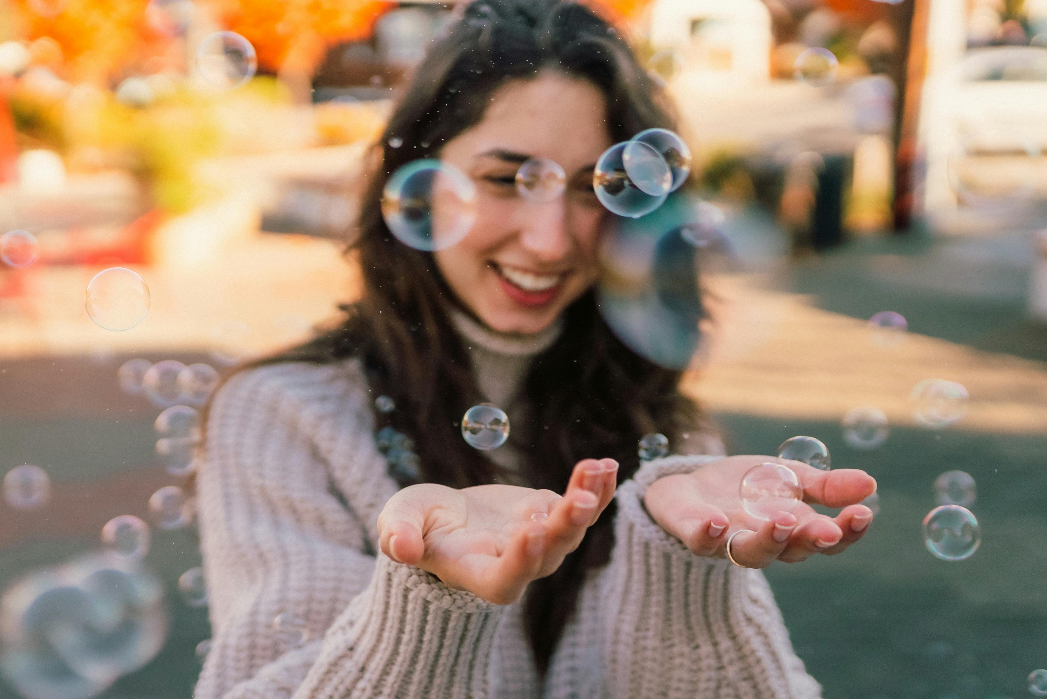 A smiling woman playfully catching bubbles in a sunny outdoor setting.