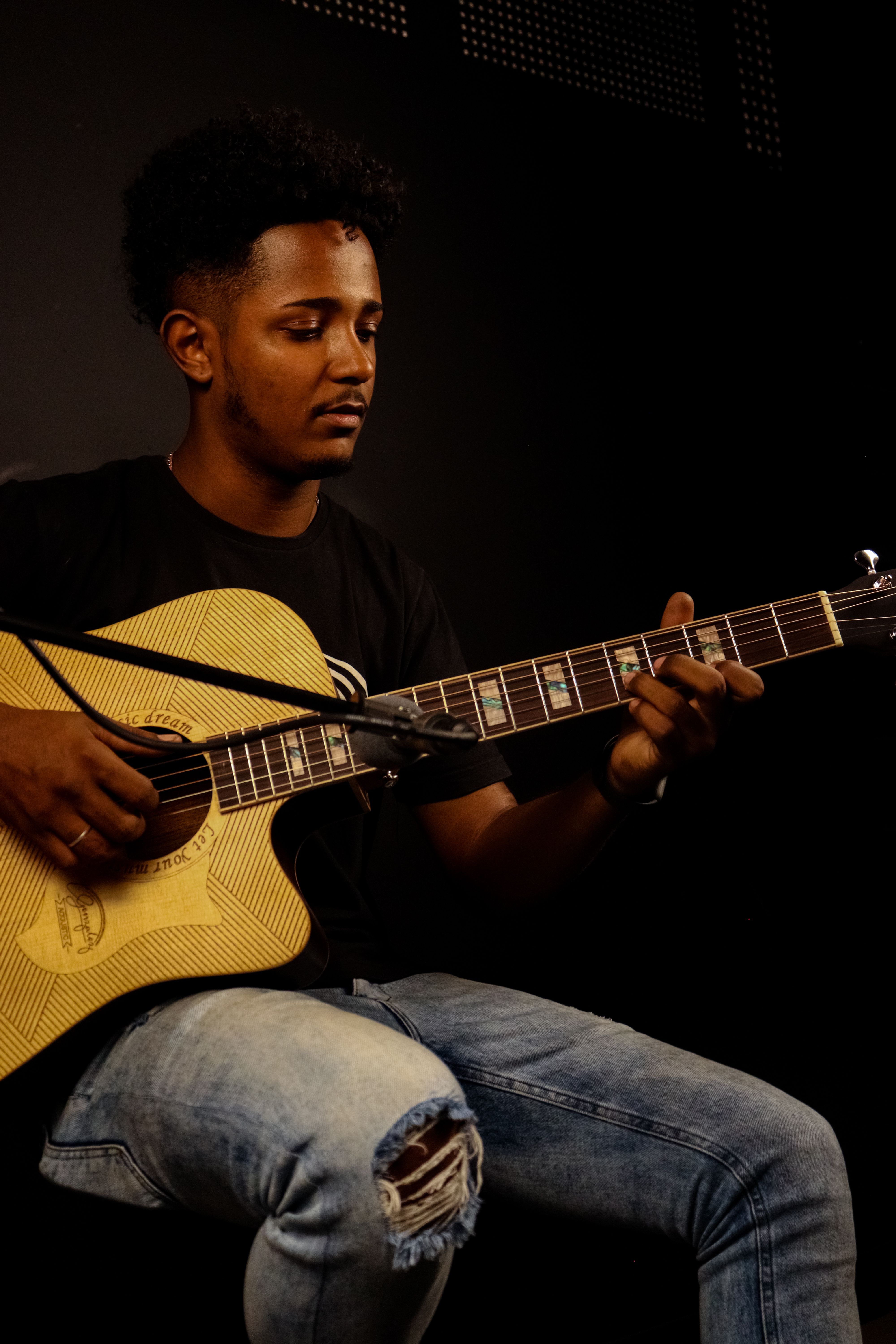 A young African American man skillfully playing an acoustic guitar in a dimly lit room.