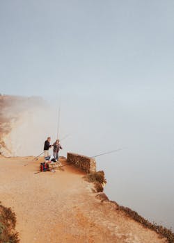 Photo by Artūras Kokorevas Two fishermen on misty cliffs in Nazaré, Portugal, capturing a peaceful seaside moment.