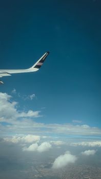 An airplane wing viewed from a window, gliding over clouds against a blue sky.