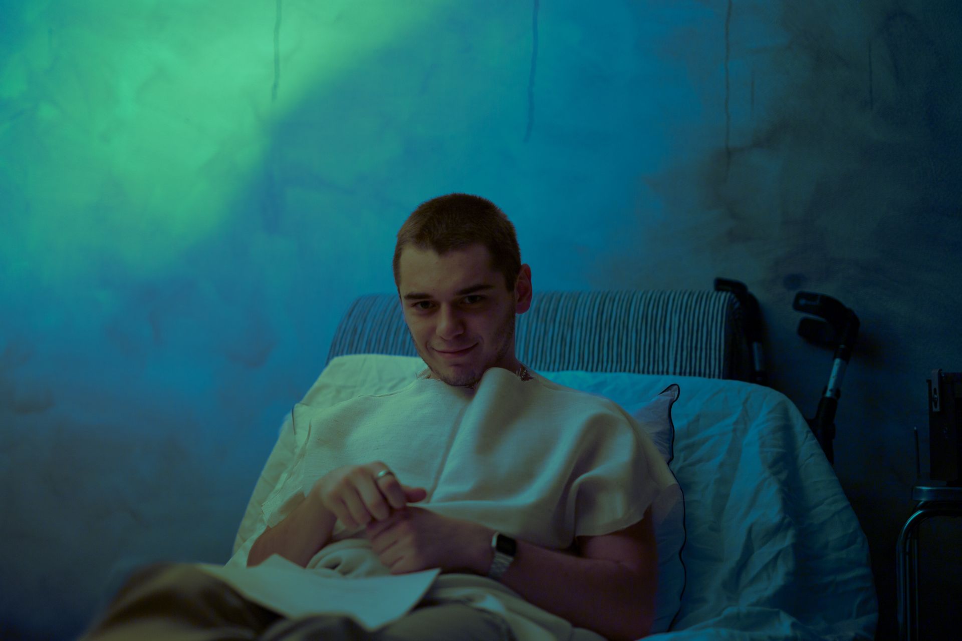 A smiling man with short hair relaxing in a hospital bed indoors.
