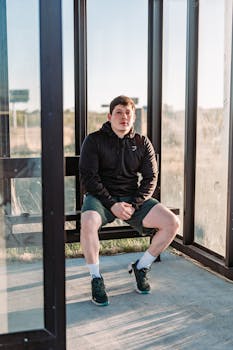 Young man in a hoodie sitting in a sunlit glass bus shelter, looking relaxed.