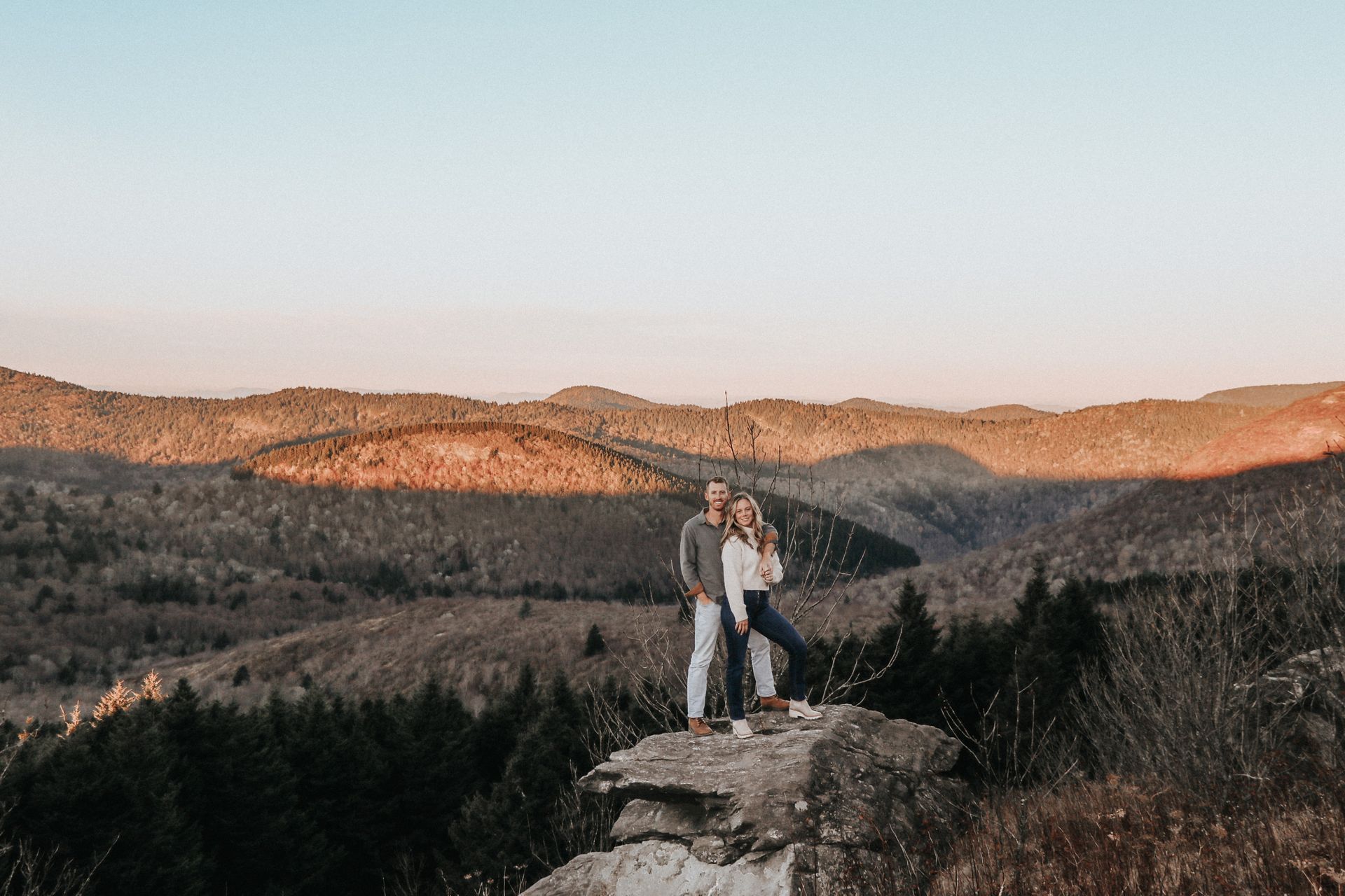 A couple stands embracing on a rock with a scenic mountain landscape as a backdrop.