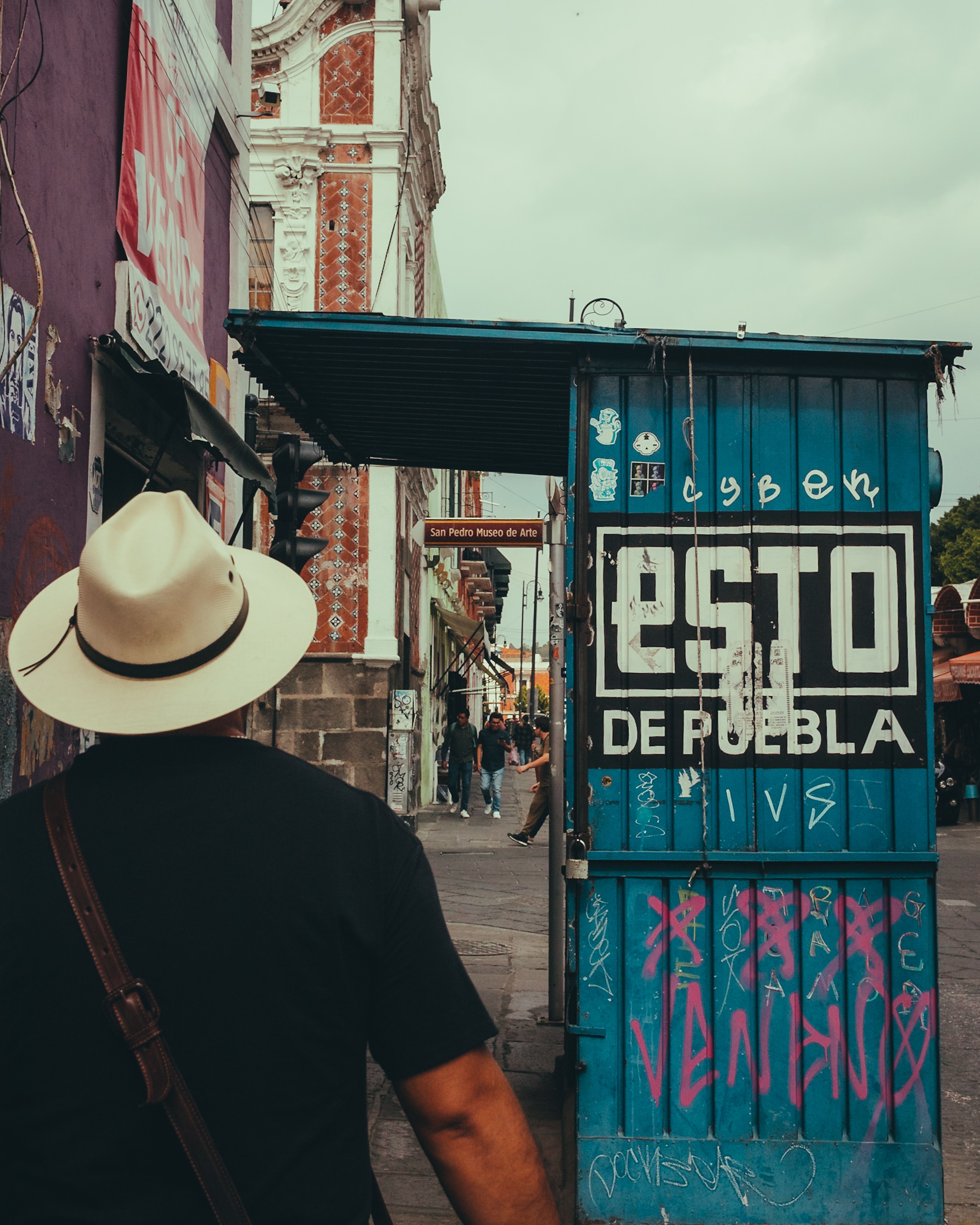 A man wearing a Panama hat walks past a colorful newsstand on a Puebla street, showcasing urban life in Mexico.