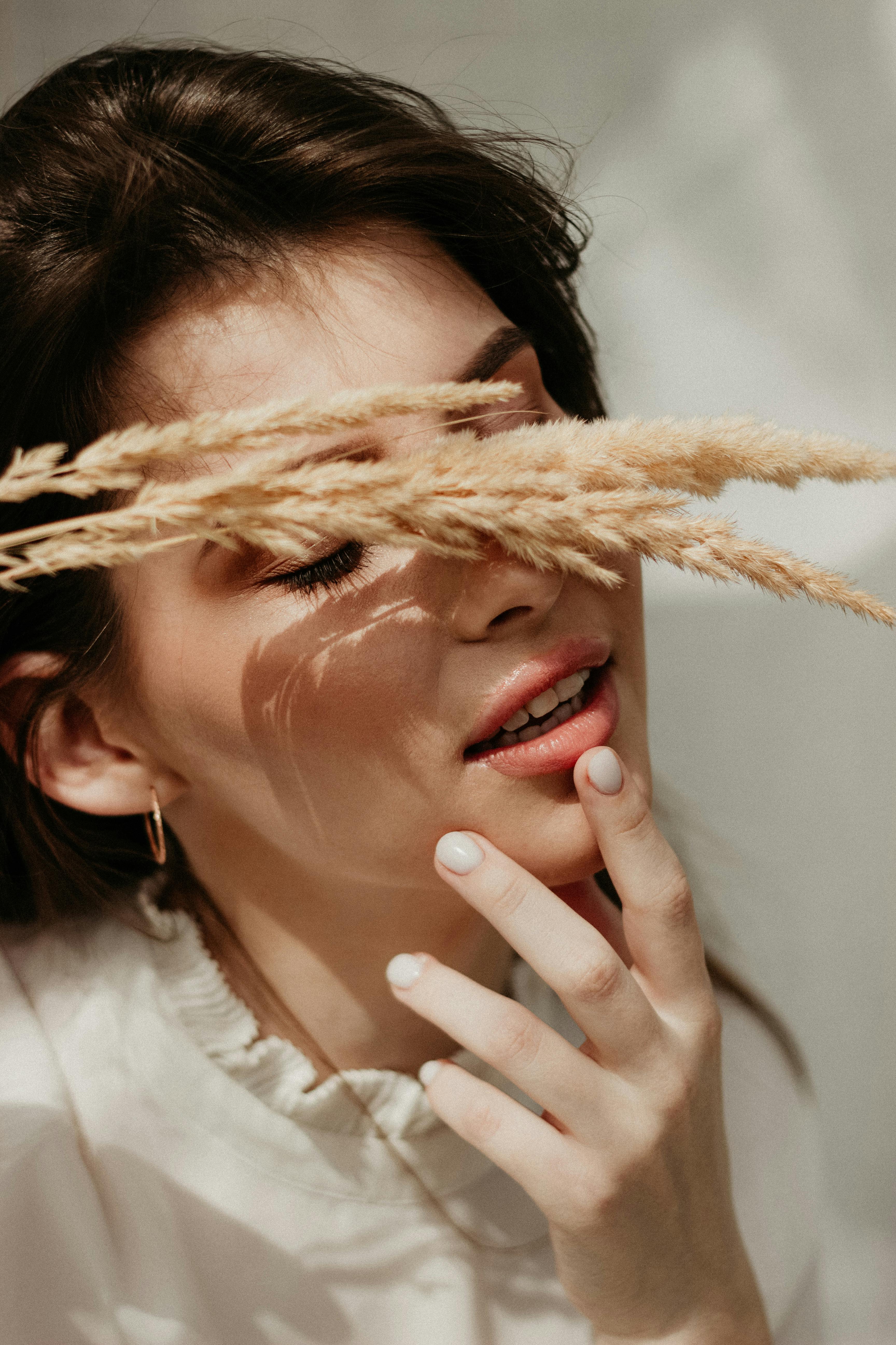 A serene portrait of a woman with her eyes closed, touching her chin with wheat grass gently brushing her face.
